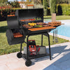 A black Royal Gourmet outdoor charcoal grill with a side ash tray and wooden lid handle, shown on a patio next to a swimming pool with food cooking on the grates.