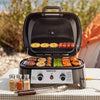 A portable outdoor Royal Gourmet gas grill with a lid, shown open on a table with food cooking inside and seasoning bottles in front. A real outdoor setting with grass and a tent is visible in the background, so this is Lifestyle rather than Product/Packaging.
