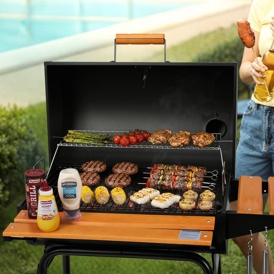A black portable outdoor Royal Gourmet grill with a wooden handle and side table, shown cooking multiple foods including burgers, steak, asparagus, tomatoes, and corn. A person is partially visible on the right holding a drink, and the grill is set in a backyard patio environment with a pool and greenery in the background.