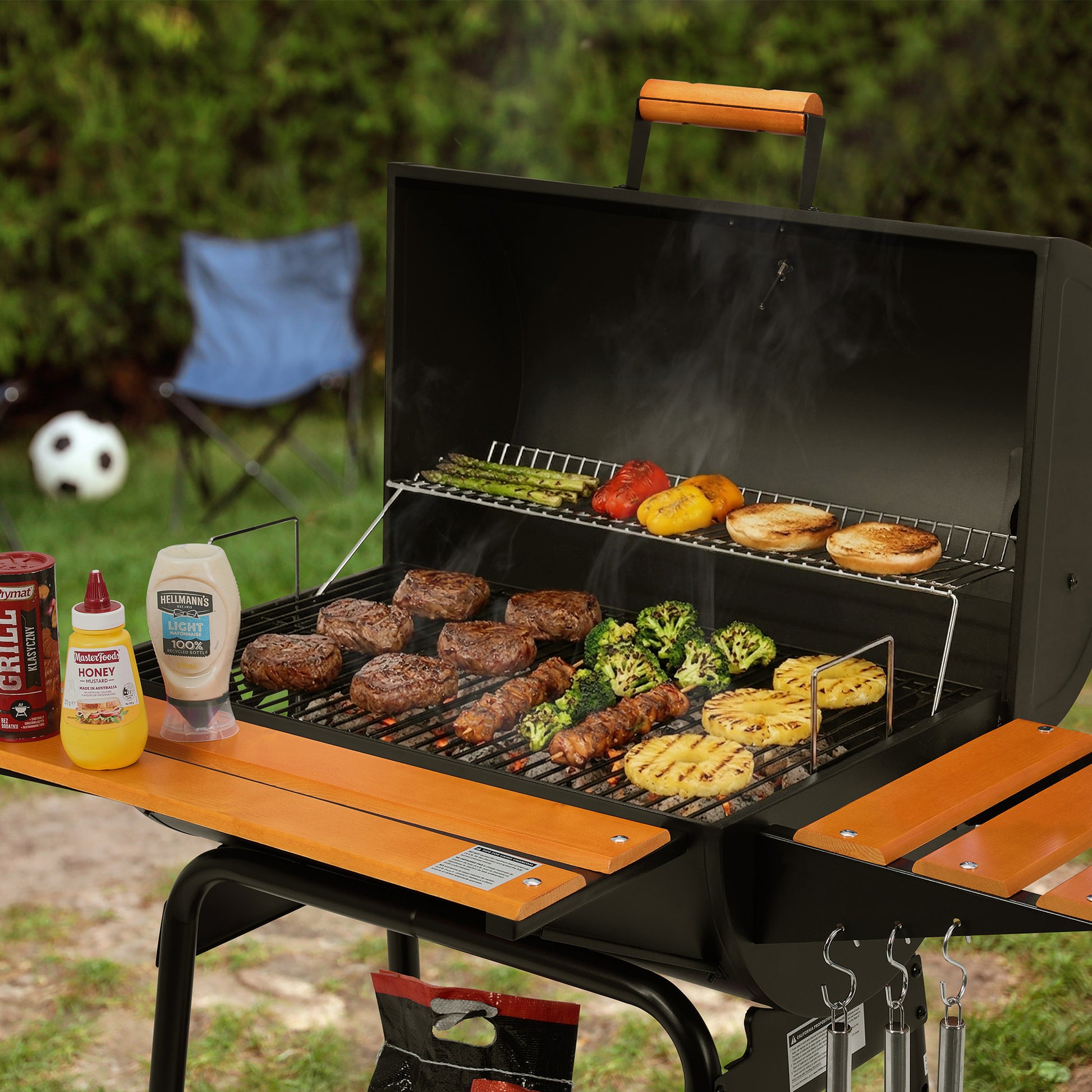 An outdoor Royal Gourmet charcoal grill with the lid open, cooking burgers, broccoli, asparagus, peppers, and buns. The grill has wooden-look side tables and handle, and is shown in a grassy backyard setting with cooking ingredients and a folding chair in the background.