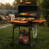 A black Royal Gourmet charcoal grill with an open lid, wooden side tables, and hanging utensils, set on a grassy backyard patio with food cooking inside and bags of charcoal underneath.