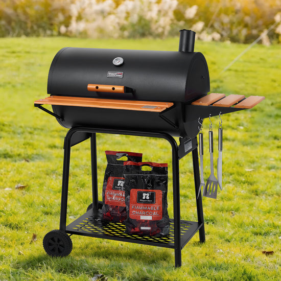 A black outdoor pellet Royal Gourmet grill on a wheeled stand with wooden slat sides, shown on a grassy lawn. Two bags of FiamaFiama hardwood charcoal sit on the lower shelf, and two hanging utensils are attached to the right side.