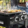 A Royal Gourmet outdoor gas grill with a flat top griddle, shown cooking meat and vegetables with visible steam rising. The black grill has two control knobs on the front and is photographed outdoors with trees and a blue tarp in the background.