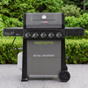 A Royal Gourmet gas grill in black and dark gray, shown outdoors on a paved surface with green hedges in the background. The grill features five control knobs, side tables on both ends, and a visible propane tank connected on the left.