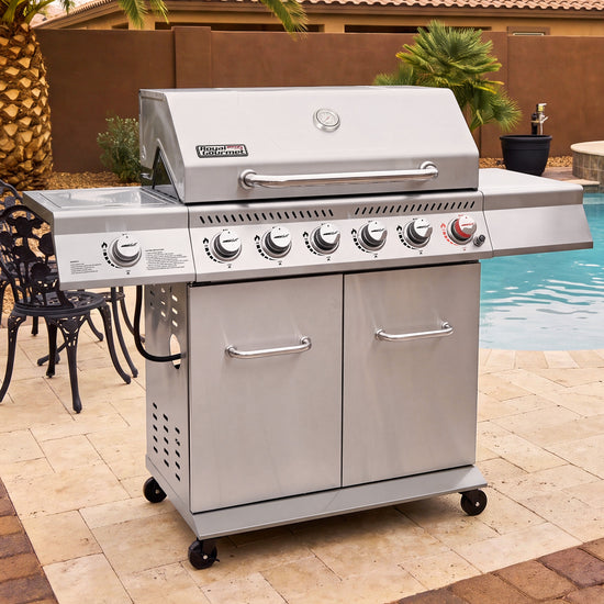 A stainless steel Royal Gourmet outdoor gas grill with side tables and dual storage cabinets, shown on a tiled patio next to a swimming pool.