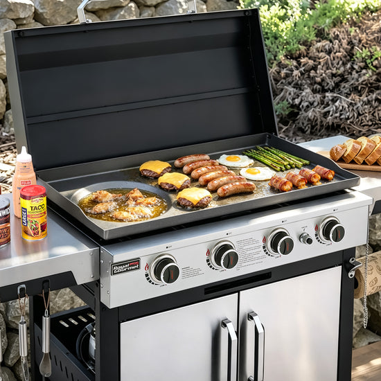 A stainless steel Royal Gourmet outdoor gas grill with the lid open, cooking sausages, eggs, asparagus, and cheese on a flat top griddle. The grill is shown outdoors on a stone patio with food props and condiments nearby.