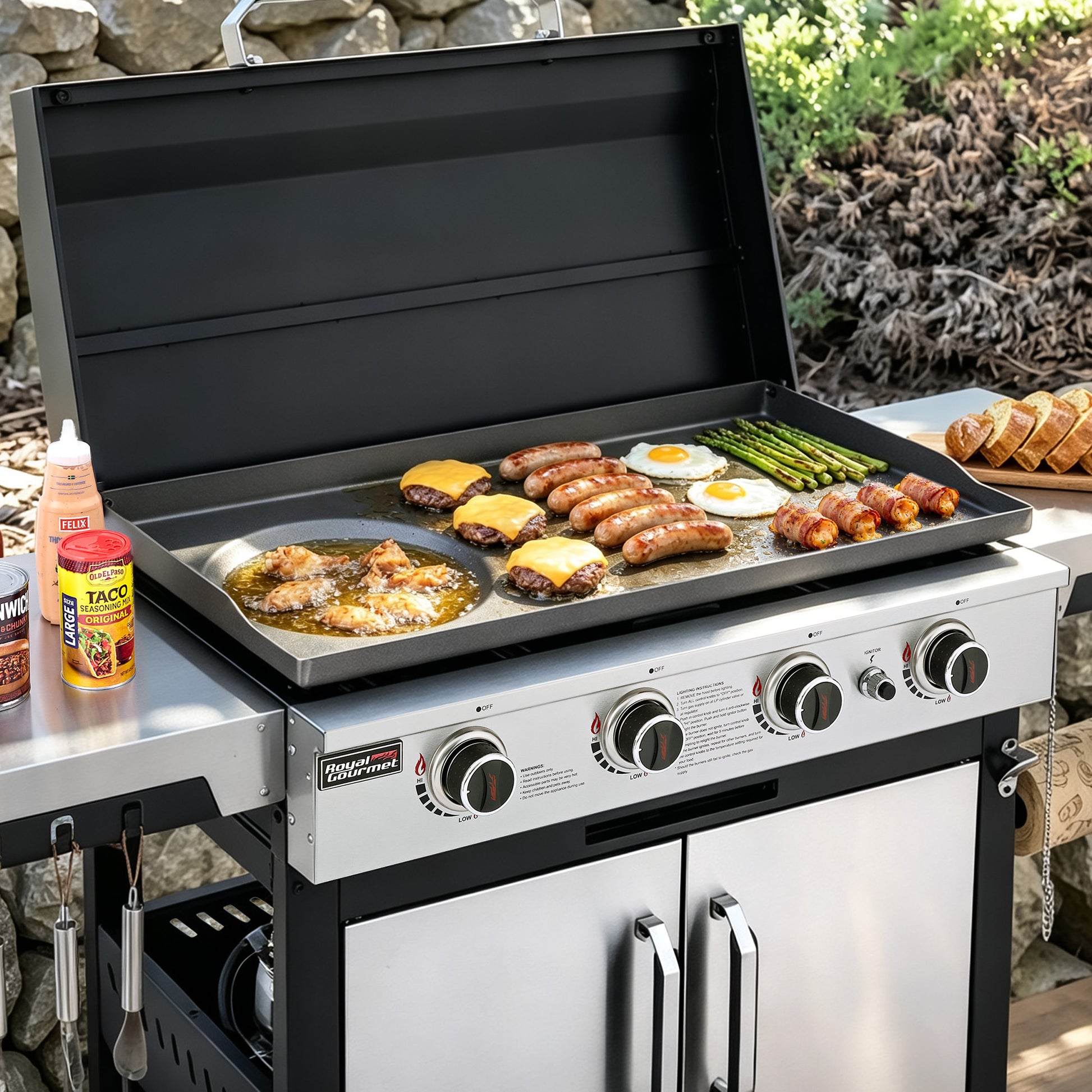 A stainless steel Royal Gourmet outdoor gas grill with the lid open, cooking sausages, eggs, asparagus, and cheese on a flat top griddle. The grill is shown outdoors on a stone patio with food props and condiments nearby.