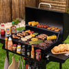 A black Royal Gourmet outdoor grill filled with food, set on grass in front of a wooden fence. Condiment bottles are arranged on the sides of the grill, and sliced bread sits on a wooden board on the right side.