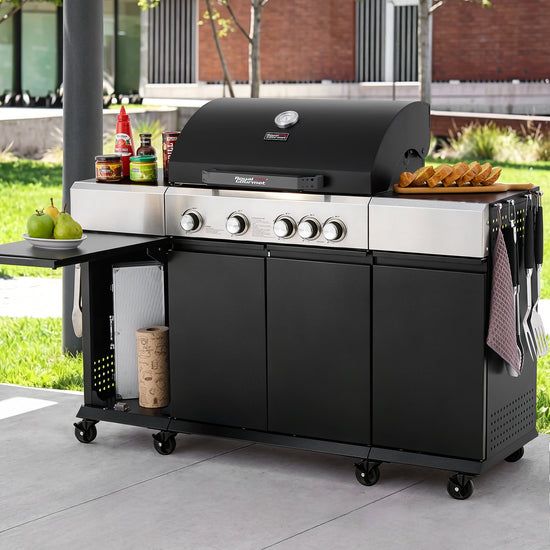 A black and stainless steel Royal Gourmet outdoor gas grill on a wheeled cart with side shelves, shown on a patio with food props and a blurred backyard background.
