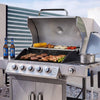 A stainless steel Royal Gourmet gas grill with four control knobs is shown outdoors on a patio, cooking steak, chicken, corn, and vegetables. Two blue beer bottles sit on the left counter, and the grill lid is open with a pot on the side.