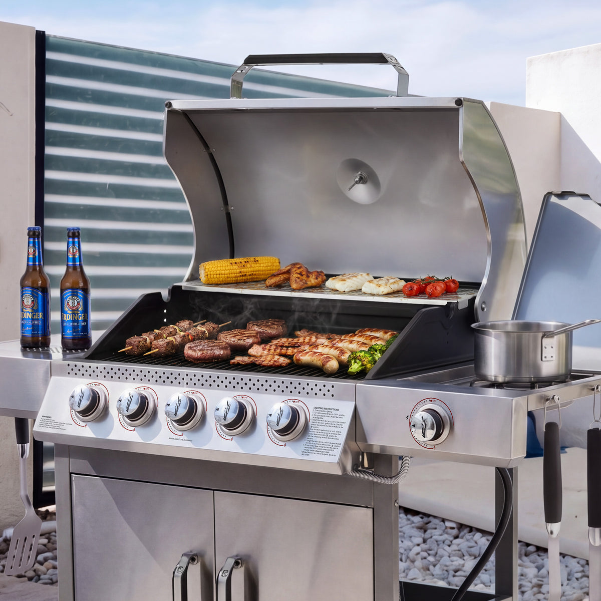 A stainless steel Royal Gourmet gas grill with four control knobs is shown outdoors on a patio, cooking steak, chicken, corn, and vegetables. Two blue beer bottles sit on the left counter, and the grill lid is open with a pot on the side.