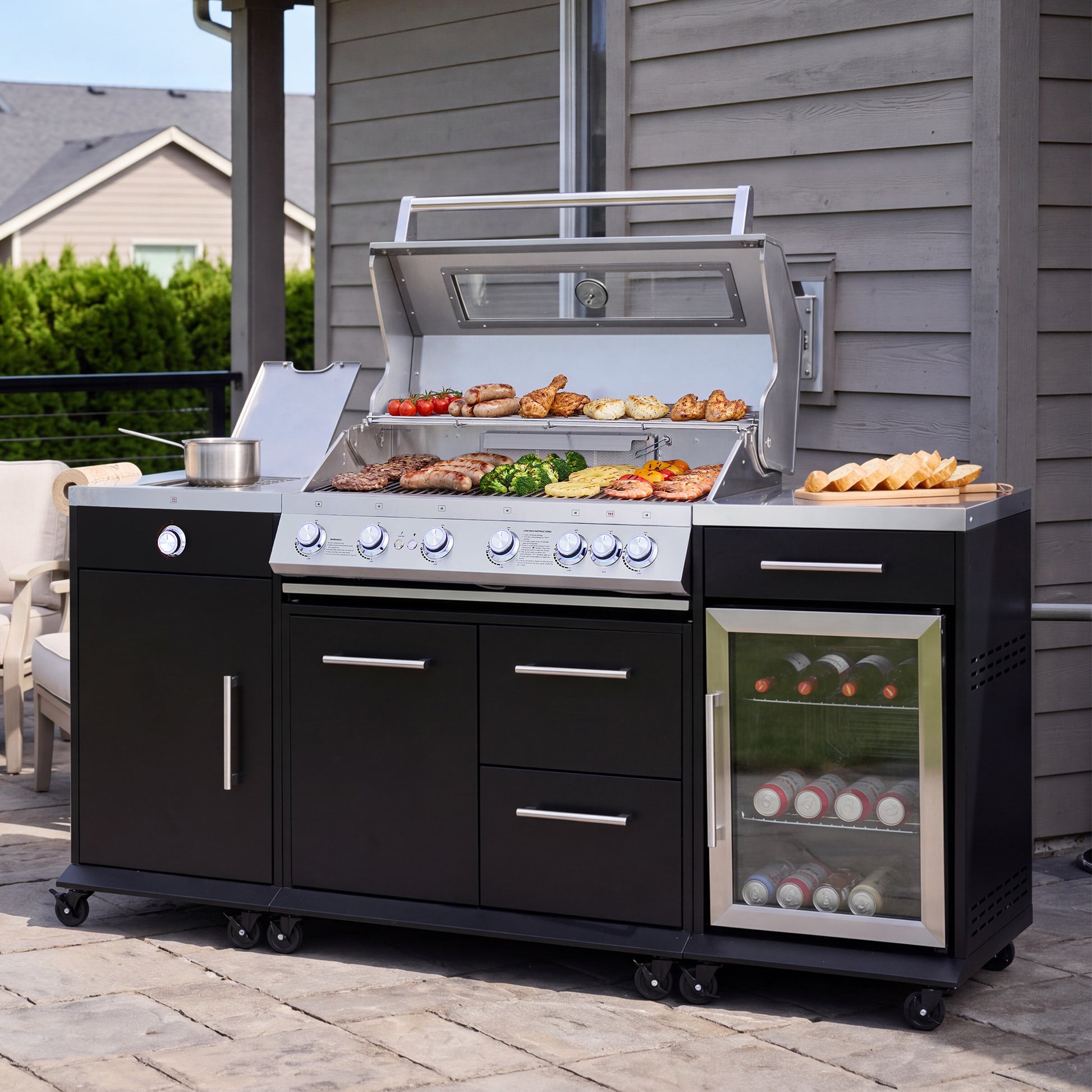 A black and stainless steel outdoor kitchen cart with a built-in Royal Gourmet  gas grill, drawers, and a glass-door wine cooler, shown on a patio with food and drinks on top.