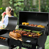 A black Royal Gourmet outdoor charcoal grill with the lid open, loaded with cooked meats, corn, peppers, and potatoes, set in a backyard with a blurred woman drinking in the background.