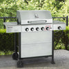 A stainless steel outdoor Royal Gourmet gas grill with a side shelf, four front control knobs, and black wheels, photographed outdoors on a patio with green hedges in the background.