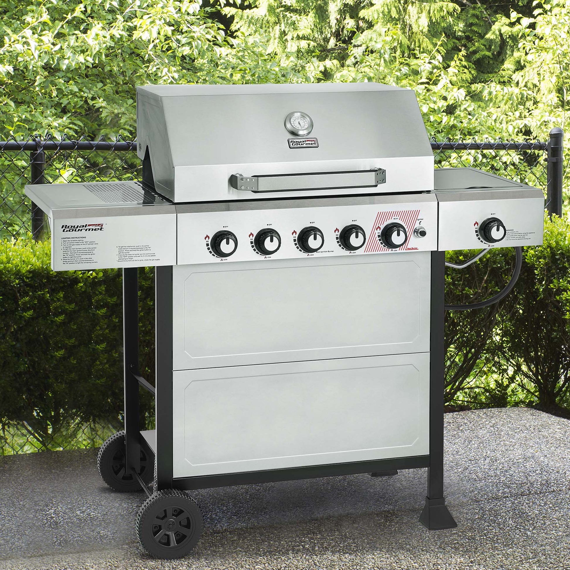 A stainless steel outdoor Royal Gourmet gas grill with a side shelf, four front control knobs, and black wheels, photographed outdoors on a patio with green hedges in the background.