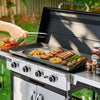 A stainless steel outdoor Royal Gourmet gas grill with a large flat top griddle, shown with food cooking on it in a backyard setting. A person's hand is visible using metal tongs to turn the food.