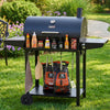 A black Royal Gourmet outdoor pellet grill on a lawn with bottles of sauces and marinades on the side table, hanging utensils, and bags of charcoal underneath. The grill lid has a visible Peloton logo.