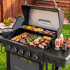 A black Royal Enfield gas grill is shown outdoors on grass with food cooking inside and condiment bottles on the side table. The grill has five control knobs on the front and an open lid with a stainless steel interior.