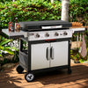 A stainless steel and black outdoor Royal Gourmet gas grill with side tables and wheels, set on a wooden deck with green foliage in the background. The grill has four control knobs and a lid-mounted thermometer.
