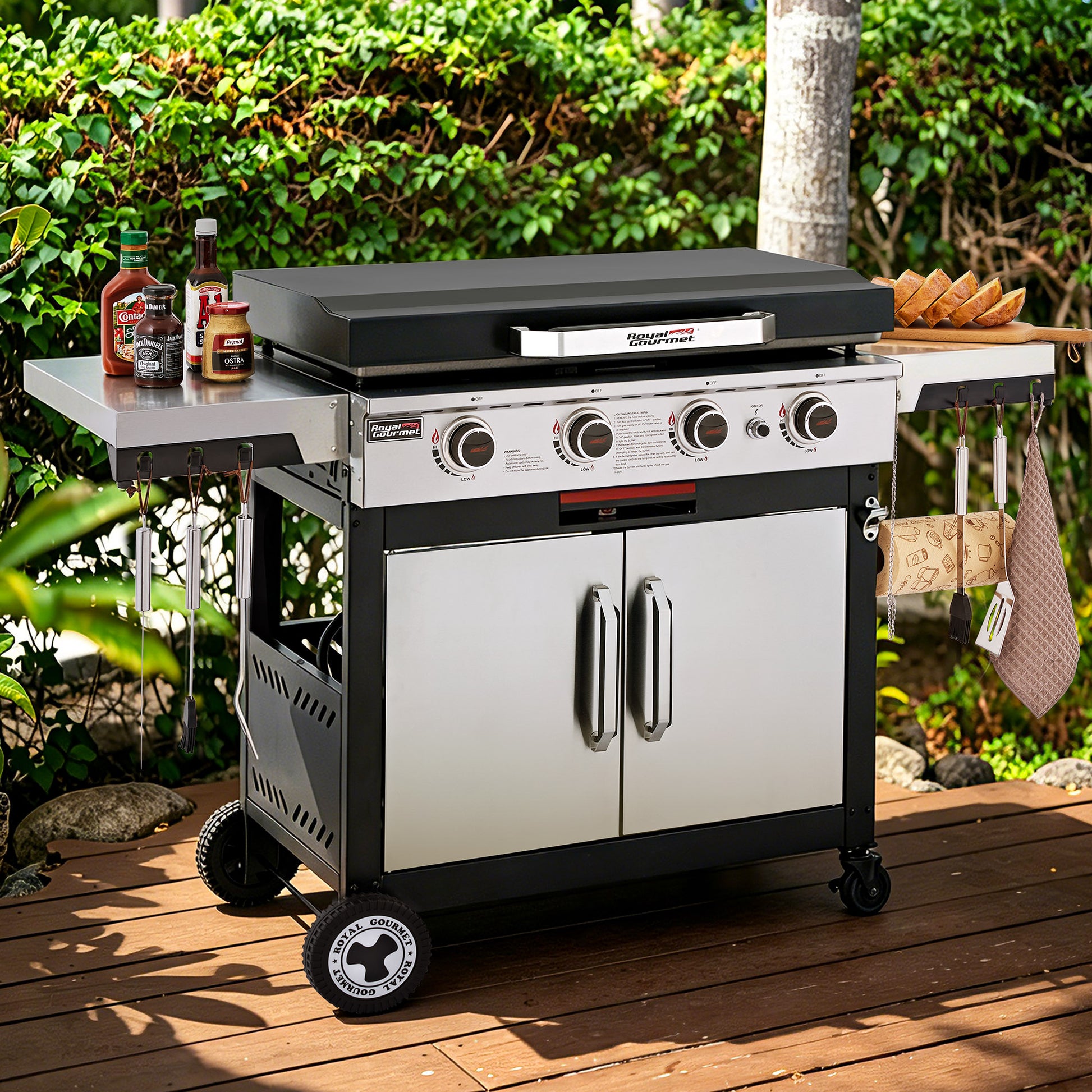 A stainless steel and black outdoor Royal Gourmet gas grill with side tables and wheels, set on a wooden deck with green foliage in the background. The grill has four control knobs and a lid-mounted thermometer.