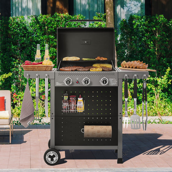 A black Royal Gourmet gas grill with side tables and hanging utensils is shown outdoors on a patio, cooking burgers, hot dogs, and vegetables, with a green garden background.