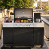 A large Royal Gourmet outdoor gas grill cart with a stainless steel top and black lower cabinets, shown open on a patio with food cooking inside and condiment bottles on the left. The setting includes trees, railings, and apartment buildings in the background.