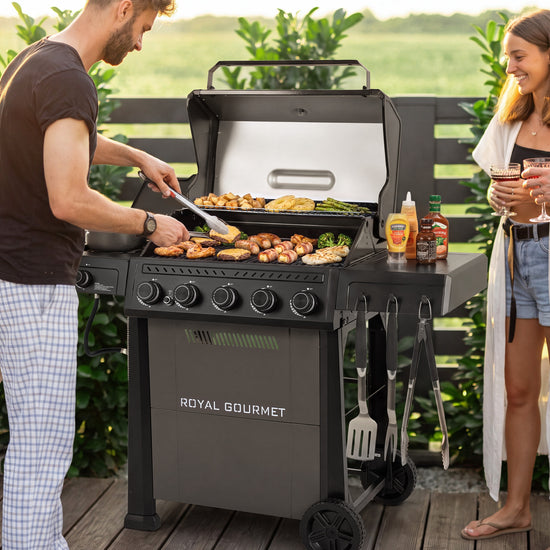 A Royal Gourmet gas grill is shown on a wooden deck outdoors, with a man cooking food inside and a woman standing nearby holding drinks. The grill is black with a stainless steel interior and side table.