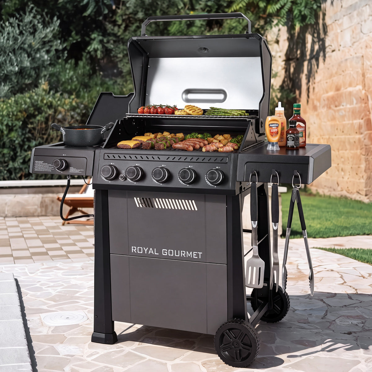 A Royal Gourmet gas grill set on a stone patio with a garden and brick wall in the background. The grill is open, showing food cooking inside, with its side table holding condiments and utensils hanging from its side hooks.