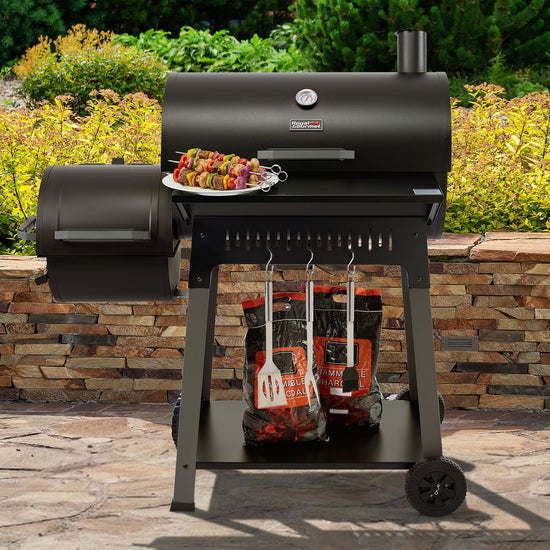 A black Royal Gourmet outdoor pellet grill on a stone patio with garden foliage in the background. A plate of skewered food sits on the grill's side table, and charcoal bags and utensils are stored underneath.