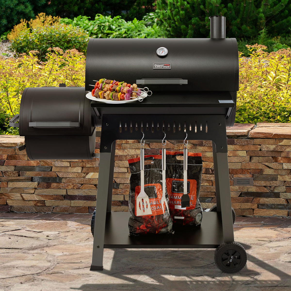 A black Royal Gourmet outdoor pellet grill on a stone patio with garden foliage in the background. A plate of skewered food sits on the grill's side table, and charcoal bags and utensils are stored underneath.