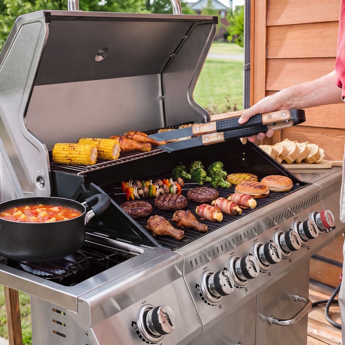 A stainless steel Royal Gourmet gas grill on a wooden deck with food cooking inside, including corn, chicken, burgers, broccoli, and skewers. A person's arm is visible holding tongs over the grill, and a pot of stew sits on the left side.