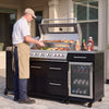 A man wearing a white shirt and tan apron is grilling food on a large black and stainless steel Royal Gourmet  outdoor gas grill with a built-in side refrigerator, set on a patio in front of a house.
