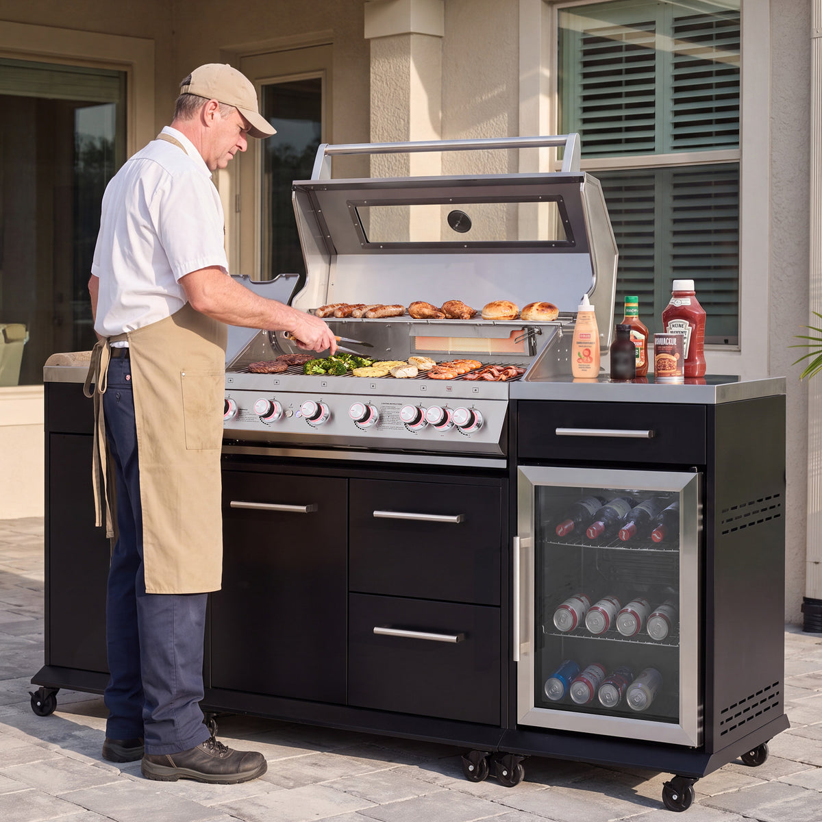 A man wearing a white shirt and tan apron is grilling food on a large black and stainless steel Royal Gourmet  outdoor gas grill with a built-in side refrigerator, set on a patio in front of a house.