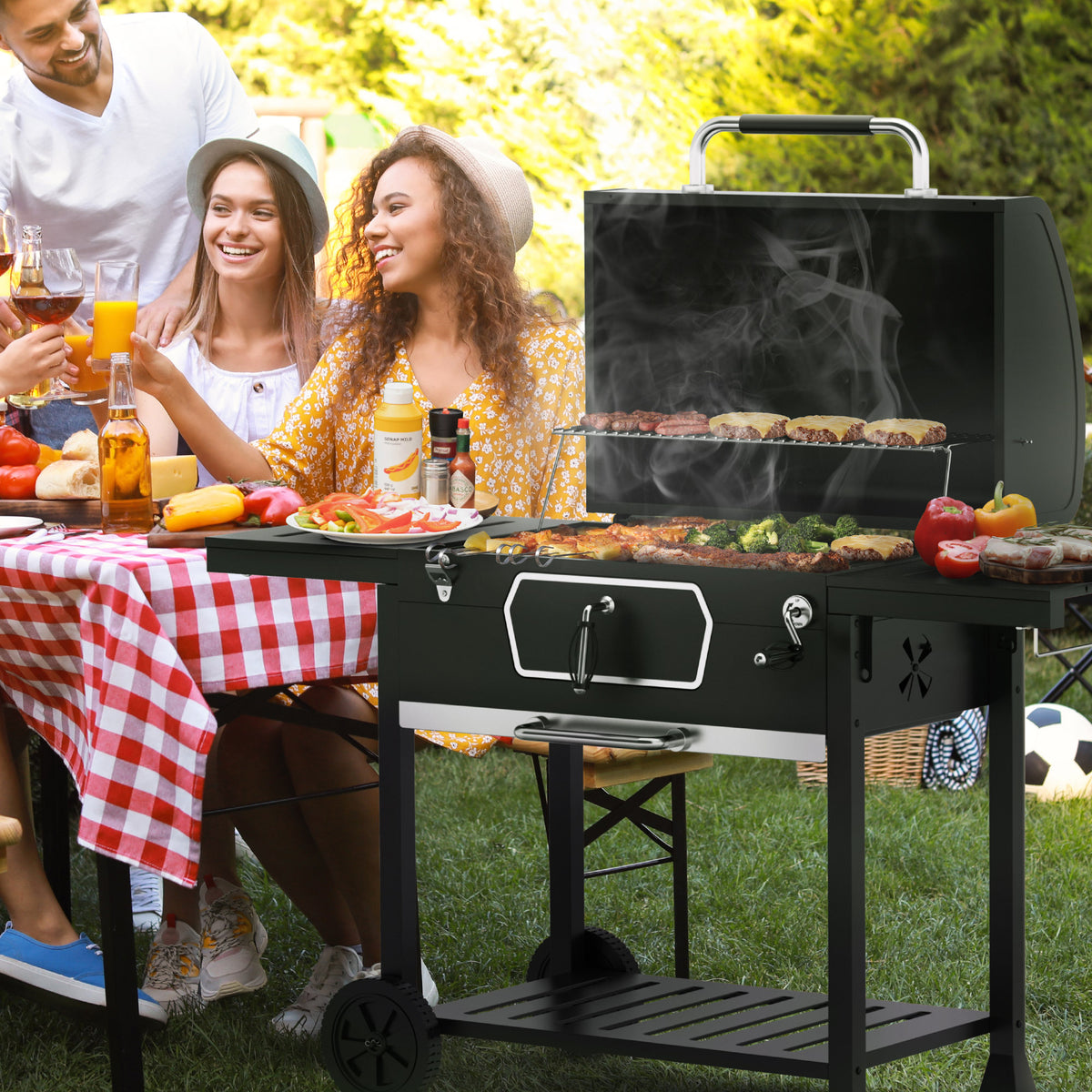 A black Royal Gourmet outdoor charcoal grill with a lid is shown on a grassy lawn during a barbecue, with people sitting at a red-and-white checkered table nearby. The grill is the product being demonstrated in a real environment with people present, so subject_type is Lifestyle.