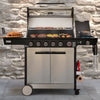 A stainless steel and black 6-burner outdoor gas grill with a glass lid, shown with food cooking on its grates and side plates. The Royal Gourmet grill is set against a rustic stone wall background.