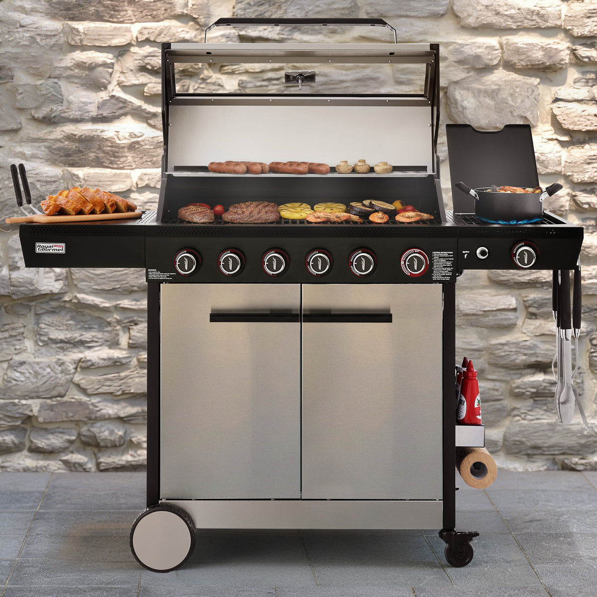 A stainless steel and black 6-burner outdoor gas grill with a glass lid, shown with food cooking on its grates and side plates. The Royal Gourmet grill is set against a rustic stone wall background.