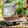 A portable Royal Gourmet electric grill with its lid open, cooking burgers, skewers, and carrots on a wooden table outdoors. A person is partially visible in the background near a table with food, set against a lush green garden backdrop.