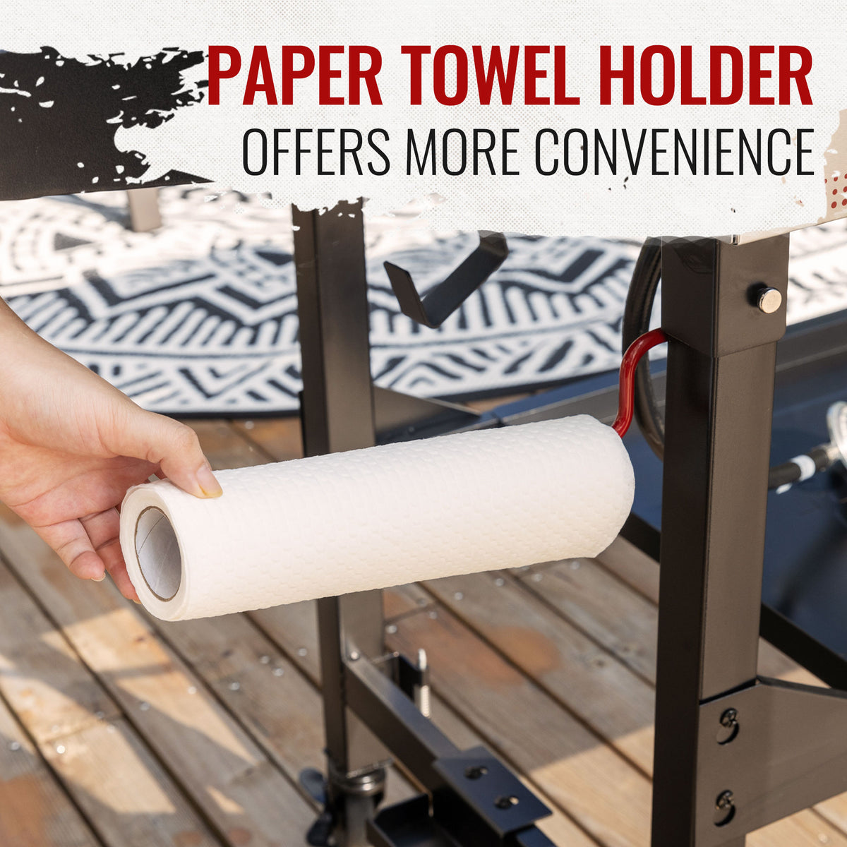 A hand holds a roll of white paper towels against a black metal stand with a red hanging loop, set on a wooden deck. Text at the top reads 'PAPER TOWEL HOLDER OFFERS MORE CONVENIENCE'.