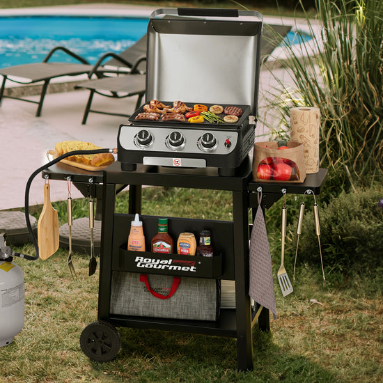 A Royal Gourmet gas grill cart with a stainless steel grill and black frame, shown outdoors on grass near a swimming pool. The cart holds cooking accessories and food, with the grill lid open and three control knobs visible.