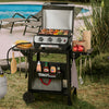 A Royal Gourmet gas grill cart with a stainless steel grill and black frame, shown outdoors on grass near a swimming pool. The cart holds cooking accessories and food, with the grill lid open and three control knobs visible.