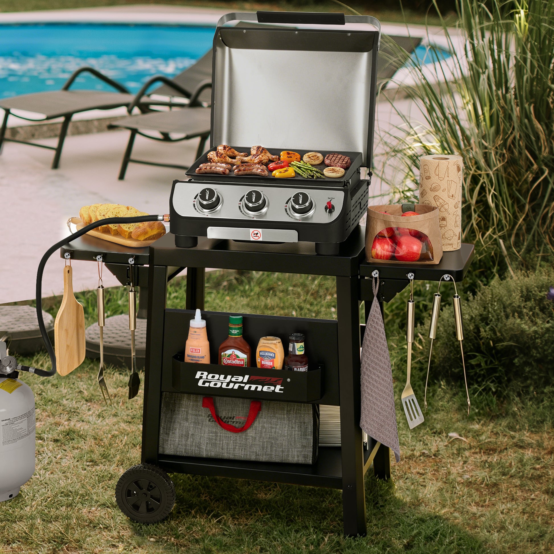 A Royal Gourmet gas grill cart with a stainless steel grill and black frame, shown outdoors on grass near a swimming pool. The cart holds cooking accessories and food, with the grill lid open and three control knobs visible.