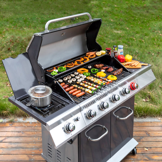 A stainless steel Royal Gourmet gas grill with the lid open, shown outdoors on a wooden deck with grass in the background. The grill is loaded with cooked meats, vegetables, skewers, and a pot on the side burner, with condiments placed on top.