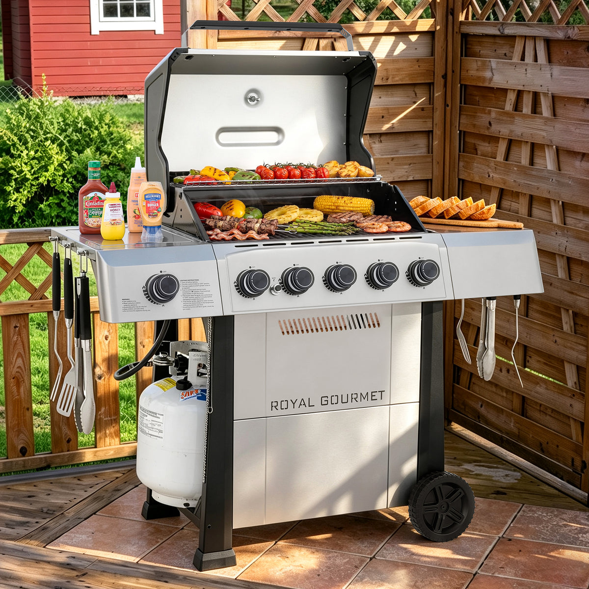 A Royal Gourmet gas grill with a stainless steel body and black wheels, shown open on an outdoor wooden deck with food cooking on the grates and condiments nearby.