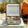 A compact outdoor electric Royal Gourmet  grill in sage green sits on a checkered tablecloth in a camping setting, with food cooking on both sides under an open lid. An orange tent is blurred in the background.