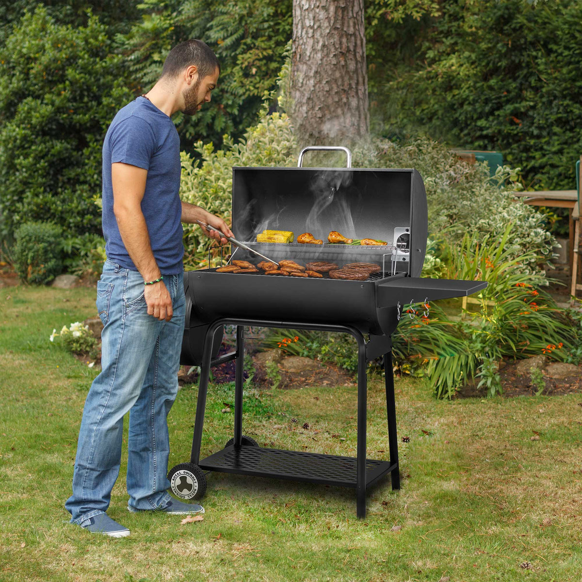 A man in a blue t-shirt and jeans grills food on a black Royal Gourmet  charcoal barbecue grill in a green backyard garden.
