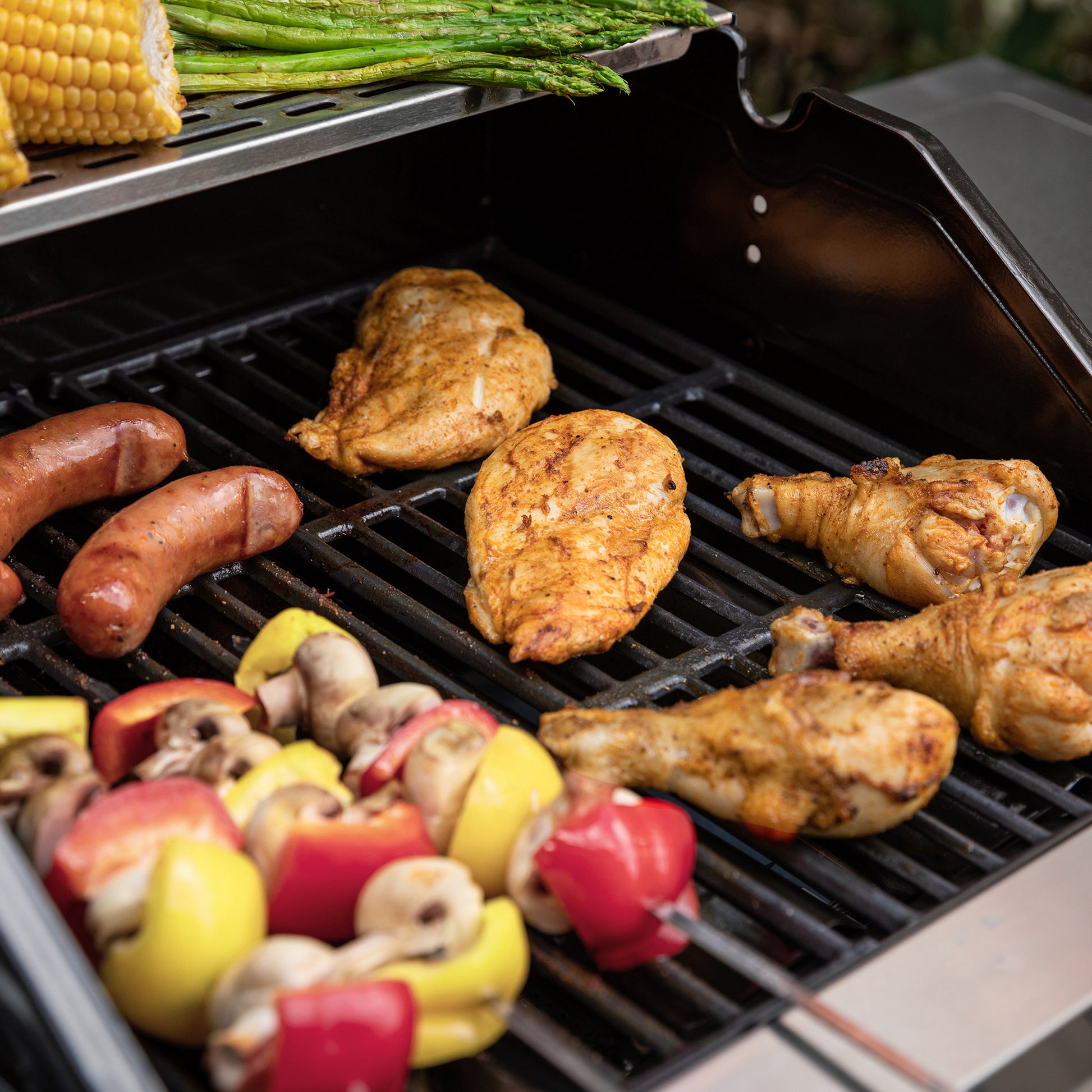 Grilled chicken pieces, sausages, mushrooms, bell peppers, and corn on a black outdoor Royal Gourmet grill with asparagus visible at the top.
