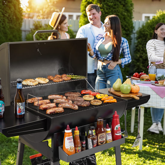 A person wearing a green shirt and blue jeans is standing behind a large Royal Gourmet  black outdoor grill, holding a metal spatula and looking toward the food on the grill. The scene is set in a backyard with a barbecue grill, bottles, and a cooler visible in the background.