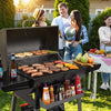 A person wearing a green shirt and blue jeans is standing behind a large Royal Gourmet  black outdoor grill, holding a metal spatula and looking toward the food on the grill. The scene is set in a backyard with a barbecue grill, bottles, and a cooler visible in the background.