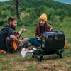 Two people sitting outdoors with a Royal Gourmet portable grill and food, surrounded by nature.