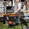 A black Royal Gourmet charcoal grill with a side table is set up on a grassy backyard patio. An older man wearing a brown apron is grilling burgers and corn, while a younger man and a woman prepare food nearby.
